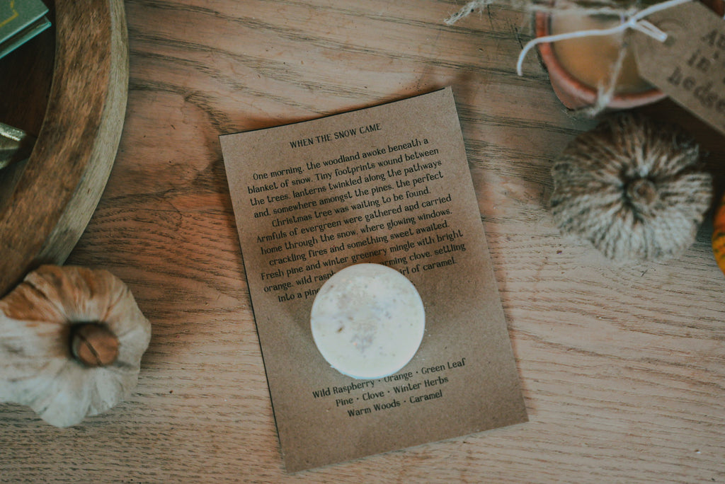 A wax melt lies on a rustic wooden surface, with a brown paper note with a poem about when the snow came. The card is styled with a dried flower, a wooden bowl, and other natural props.